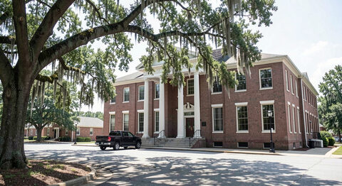 A quiet county courthouse square in Mississippi at midday, a low brick building with tall windows and a Confederate-free plain portico, live oaks and a parked pickup.